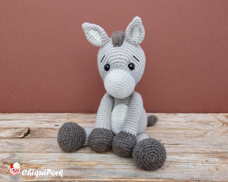 Puede incluir: Un juguete de peluche de burro de crochet gris con ojos negros y nariz blanca. El burro est&aacute; sentado sobre una superficie de madera.