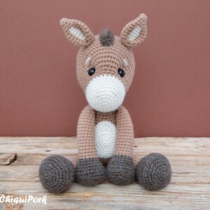 Puede incluir: Un juguete de burro de crochet marr&oacute;n y gris sentado en una superficie de madera. El burro tiene detalles blancos en la cara y las pezu&ntilde;as.