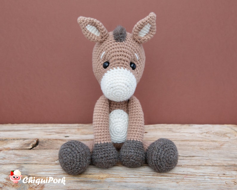 Puede incluir: Un juguete de burro de crochet marr&oacute;n y gris sentado en una superficie de madera. El burro tiene detalles blancos en la cara y las pezu&ntilde;as.
