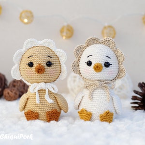 May include: Two crocheted ducklings with black eyes, orange beaks, and pink cheeks. The left duckling is tan with a white bonnet, and the right duckling is white with a tan bonnet. Both have orange feet and are tied with matching yarn bows.