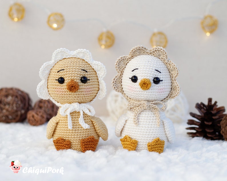 May include: Two crocheted ducklings with black eyes, orange beaks, and pink cheeks. The left duckling is tan with a white bonnet, and the right duckling is white with a tan bonnet. Both have orange feet and are tied with matching yarn bows.