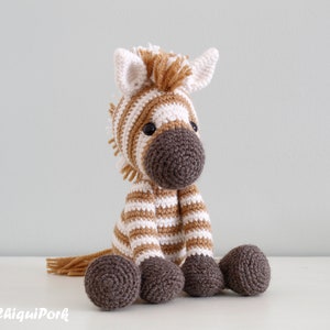 May include: A crocheted zebra stuffed animal with brown and white stripes. The zebra has a brown nose and black eyes. The zebra is sitting on a white surface.