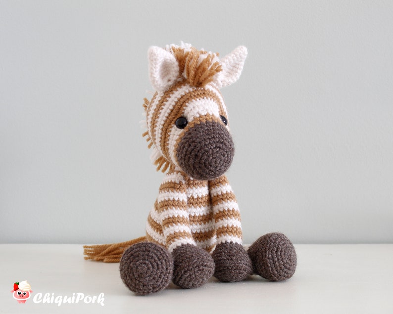 May include: A crocheted zebra stuffed animal with brown and white stripes. The zebra has a brown nose and black eyes. The zebra is sitting on a white surface.