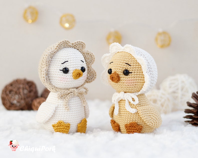 May include: Two crocheted ducklings with black bead eyes and pink cheeks. The left duckling is white with a beige bonnet and yellow feet. The right duckling is light brown with a white bonnet and brown feet. Both have small orange beaks.