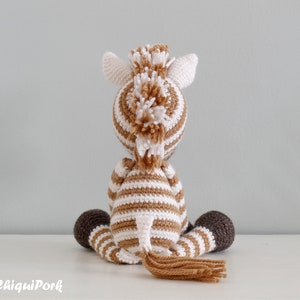 May include: A brown and white striped crocheted zebra toy with a brown tail and brown hooves. The zebra is sitting on a white surface.