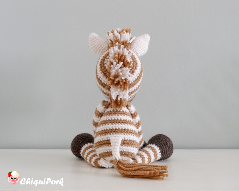 May include: A brown and white striped crocheted zebra toy with a brown tail and brown hooves. The zebra is sitting on a white surface.