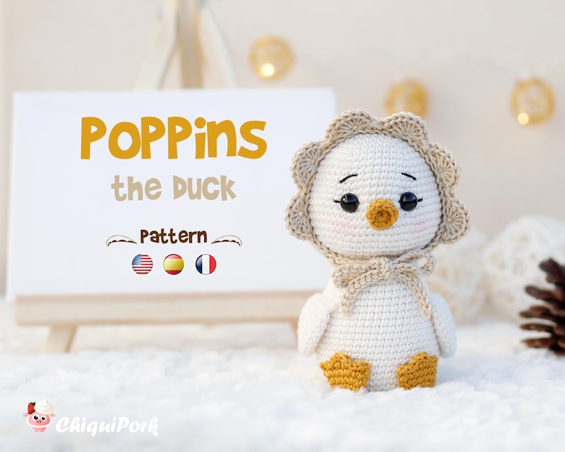May include: A white crocheted duck doll with a beige scalloped bonnet, orange beak, and orange feet. Beside it, a white sign displays the text 'POPPins the DUCK Pattern' with American, Spanish, and French flag icons and a small pig logo.