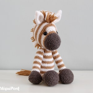 May include: A crocheted zebra stuffed animal with brown and white stripes. The zebra has a brown nose and black eyes. The zebra is sitting on a white surface.