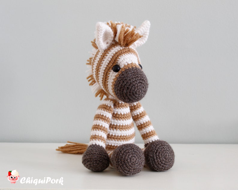 May include: A crocheted zebra stuffed animal with brown and white stripes. The zebra has a brown nose and black eyes. The zebra is sitting on a white surface.