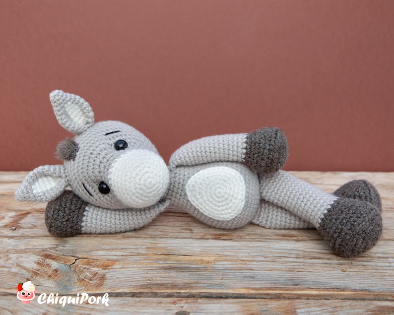 Puede incluir: Un juguete de peluche de burro gris tejido a crochet con pezu&ntilde;as marrones y una nariz blanca. El burro est&aacute; acostado sobre una superficie de madera.
