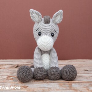 Puede incluir: Un juguete de peluche de burro de crochet gris y blanco sentado en una superficie de madera. El burro tiene una larga melena y cola grises, y sus ojos son negros con pupilas blancas.