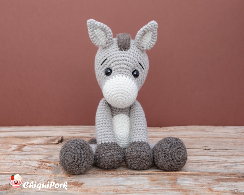 Puede incluir: Un juguete de peluche de burro de crochet gris y blanco sentado en una superficie de madera. El burro tiene una larga melena y cola grises, y sus ojos son negros con pupilas blancas.