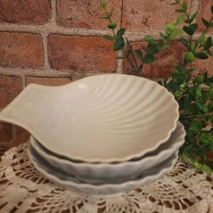 May include: A stack of three white ceramic seashell-shaped dishes. The top dish has a handle and a ridged design. The dishes are stacked on a white lace doily, with a brick wall background and green foliage.