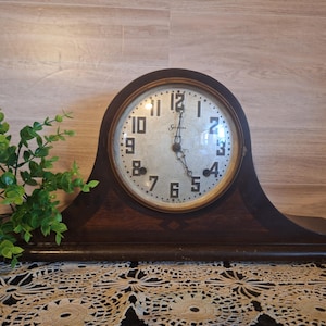 May include: A vintage wooden mantel clock with a round face. The clock face has black numerals and hands, with the brand name "Sessions" at the top. The clock is dark brown and sits on a shelf.
