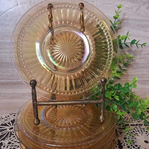 May include: A stack of amber-colored glass plates with a decorative beaded rim and a starburst design in the center. The plates are displayed on a vintage metal plate stand, with a few plates on top. Greenery is in the background.