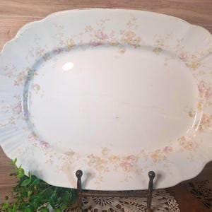 May include: Oval, white serving platter with a scalloped edge and floral pattern in pink, yellow, and green. The vintage platter is displayed on a metal stand, showcasing its elegant design. A classic piece for serving food.