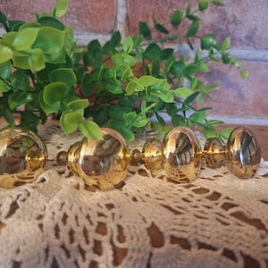 May include: Four gold-toned cabinet knobs with a round, slightly domed shape. The knobs are arranged in a row on a white crocheted doily.