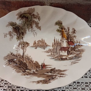 May include: A white ceramic serving dish with a brown and cream colored design of a countryside scene with a windmill, a house, and trees. The dish has a scalloped edge.