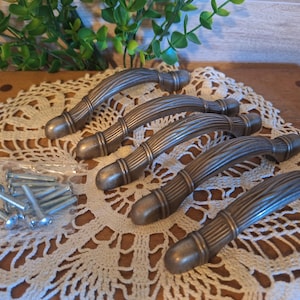 May include: Six antique bronze cabinet pulls with screws. The pulls have a curved, ribbed design with rounded ends. They are arranged on a white lace doily, with greenery in the background. Hardware for home decor.