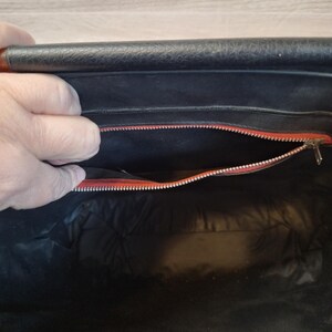May include: Open view of a black handbag interior with a red zipper and lining. The bag's interior is visible, revealing a spacious compartment. The zipper is silver, and the lining is a vibrant red. The bag appears to be made of leather.