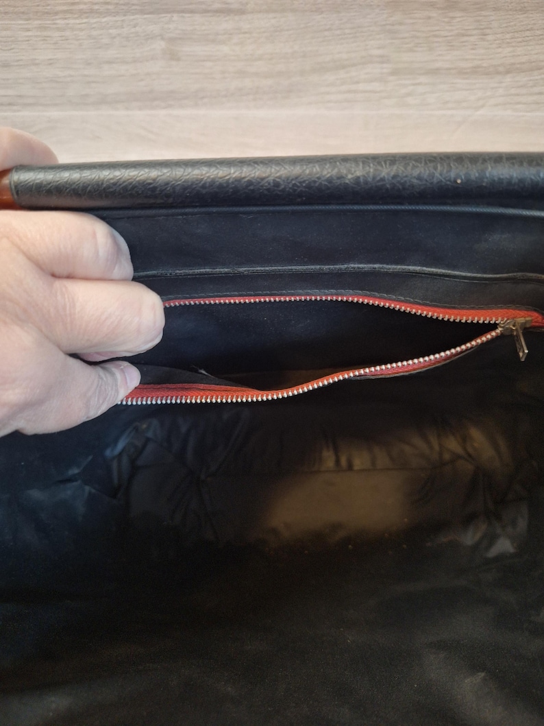 May include: Open view of a black handbag interior with a red zipper and lining. The bag's interior is visible, revealing a spacious compartment. The zipper is silver, and the lining is a vibrant red. The bag appears to be made of leather.
