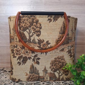 May include: A beige tote bag with a brown landscape tapestry design. The bag has a black handle and two curved wooden handles. The tapestry depicts trees, buildings, and floral elements.