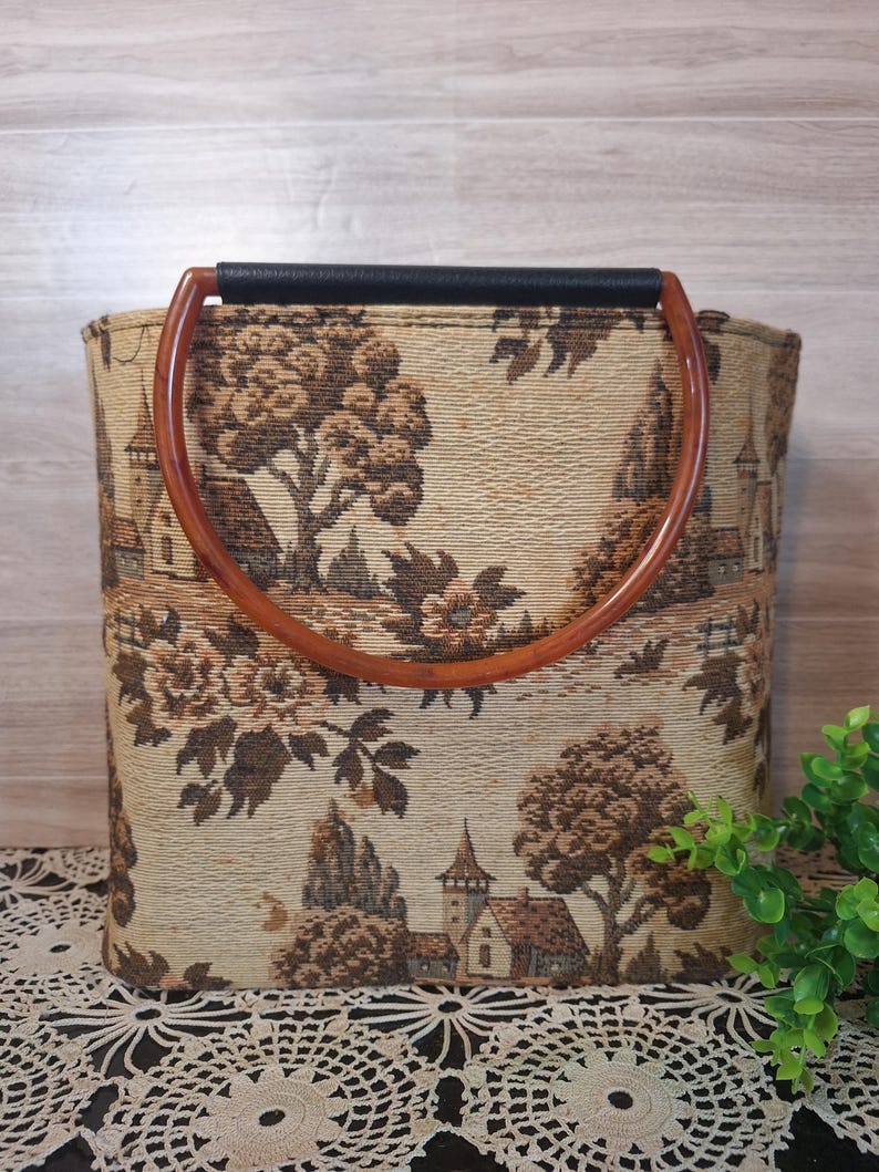 May include: A beige tote bag with a brown landscape tapestry design. The bag has a black handle and two curved wooden handles. The tapestry depicts trees, buildings, and floral elements.
