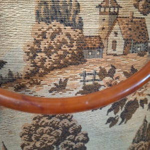 May include: Close-up of a vintage tapestry featuring a pastoral landscape with trees, buildings, and a wooden handle. The tapestry has a beige background with brown and black details, and a curved, brown handle.