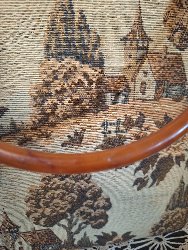 May include: Close-up of a vintage tapestry featuring a pastoral landscape with trees, buildings, and a wooden handle. The tapestry has a beige background with brown and black details, and a curved, brown handle.