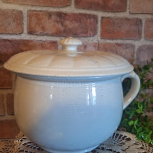 May include: A vintage, light blue and cream-colored ceramic chamber pot with a lid. The pot has a rounded shape with a handle and a decorative lid. The background features a brick wall and a lace doily.