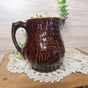 May include: A brown ceramic pitcher with a textured, tree-bark-like exterior. The pitcher features a detailed relief of a cabin and trees. It has a curved handle and a spout for pouring. The pitcher is displayed on a wooden surface with a lace doily.