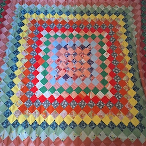May include: A colorful handmade quilt with a diamond pattern. The quilt features a variety of fabrics in shades of red, orange, yellow, green, blue, and pink. The quilt's design is a series of concentric squares, creating a vibrant and eye-catching textile.