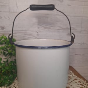May include: A white enamel bucket featuring a dark blue rim and a metal handle with a black grip. The cylindrical container has a smooth finish and a sturdy construction, designed for utility with a simple, functional aesthetic and clear lines.