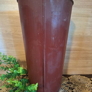 May include: A tall, vintage, burgundy-colored metal bucket with a slightly flared shape. The bucket has a seam down the front and a dark rim. The background includes a wooden surface and a sprig of green leaves.