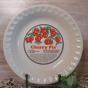 May include: White ceramic pie plate with a scalloped edge, featuring a cherry pie recipe in the center. The recipe includes ingredients like sugar, almond extract, and cherries. The plate is displayed on a decorative metal stand.