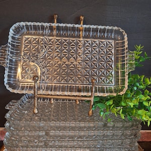 May include: A stack of clear glass serving trays with a diamond pattern, displayed on a decorative metal stand. The trays have a rectangular shape with handles and a scalloped edge. A sprig of green foliage is in the background.