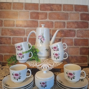 May include: A vintage tea set with a floral design. The set includes a teapot, sugar bowl, creamer, and six cups and saucers. The cups and saucers are decorated with pink roses and green leaves. The teapot has a matching floral design and a lid with a knob.