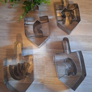 May include: Four stainless steel dreidel cookie cutters. Each cutter has a distinct shape, with a handle at the top and a stylized letter inside. The cutters are arranged on a wooden surface, with a sprig of greenery in the background.