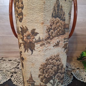 May include: A vintage cylindrical tote bag with a tapestry design featuring a landscape scene in shades of brown and beige. It has two curved, brown plastic handles. The bag's fabric has a woven texture.