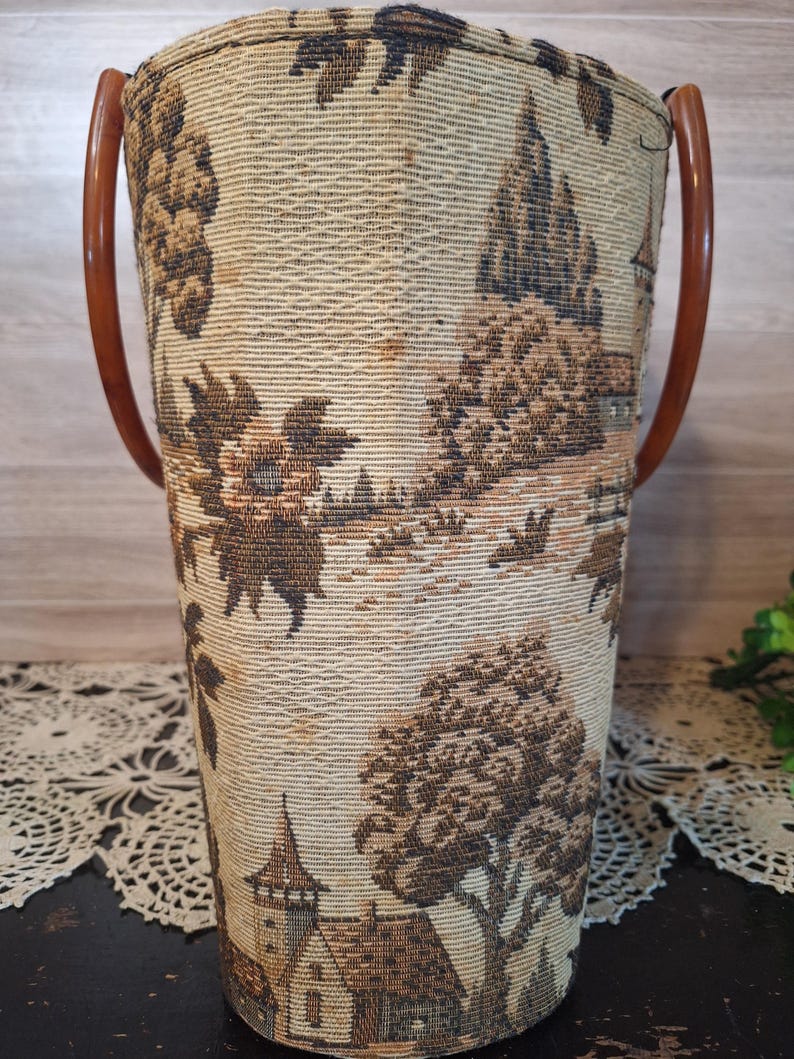 May include: A vintage cylindrical tote bag with a tapestry design featuring a landscape scene in shades of brown and beige. It has two curved, brown plastic handles. The bag's fabric has a woven texture.