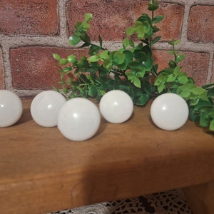 May include: Six white, spherical decorative knobs are arranged on a wooden surface, with green foliage in the background. The knobs are evenly spaced and have a glossy finish. The background features a brick wall.