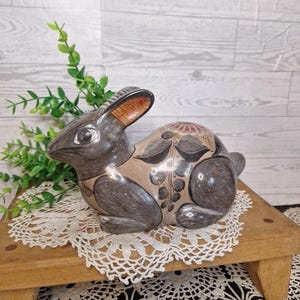 May include: A ceramic rabbit figurine featuring a smooth, polished finish in shades of dark gray and tan. The body is decorated with hand-painted black floral and leaf motifs, while the ears display a warm brown interior tone and dark outer edges.