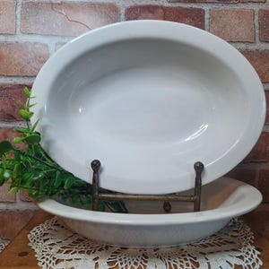 May include: A white oval ceramic serving dish with a smooth, glossy finish. The dish is displayed on a dark metal stand, with a small sprig of green artificial leaves tucked behind the base of the stand, resting on a white lace doily.