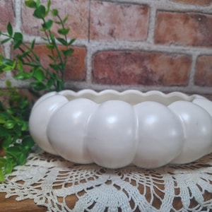 May include: A low, round, off-white ceramic bowl with a scalloped, pumpkin-like exterior featuring segmented, rounded sides. The bowl has a smooth, glossy finish and a wide, open top, presenting a simple, functional design for holding various small items.