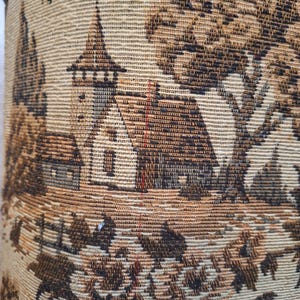 May include: Close-up of a woven fabric featuring a detailed landscape scene. The design includes a church with a steeple, trees, and foliage in shades of brown and beige. The fabric has a textured appearance.