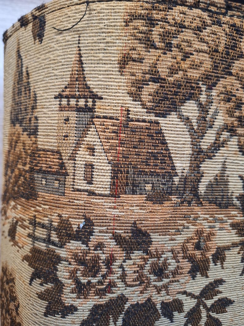 May include: Close-up of a woven fabric featuring a detailed landscape scene. The design includes a church with a steeple, trees, and foliage in shades of brown and beige. The fabric has a textured appearance.