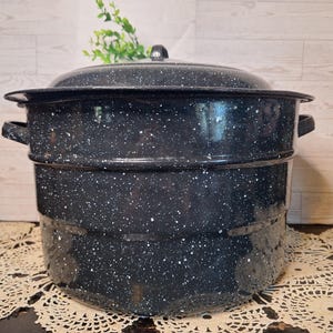 May include: A large, round, black enamel stockpot with a white speckled pattern. The pot features two side handles, a tiered cylindrical body, and a matching lid with a small central knob handle on top. The surface has a glossy, reflective finish.