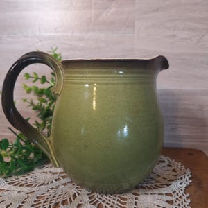 May include: A handmade, green ceramic pitcher with a dark brown rim and handle. The pitcher has a rounded body and a spout for pouring. The handle is a darker shade of brown. The pitcher is sitting on a wooden surface.