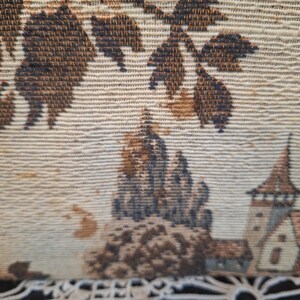 May include: Close-up of a tapestry with a detailed landscape scene. The tapestry features a beige background with brown and dark blue accents. The design includes trees, a castle, and a church.