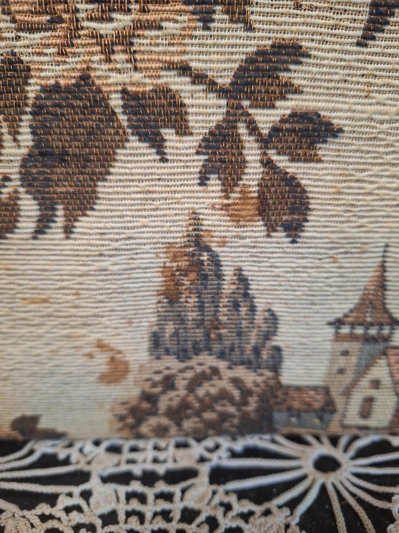 May include: Close-up of a tapestry with a detailed landscape scene. The tapestry features a beige background with brown and dark blue accents. The design includes trees, a castle, and a church.